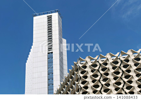 Modern white high rise skyscraper next to a geometric concrete patterned building under clear blue sky 141088433
