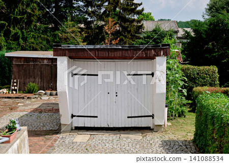 White wooden double garage doors with black hinges in a green backyard garden setting White wooden double garage doors with black hinges in a green backyard garden setting 141088534