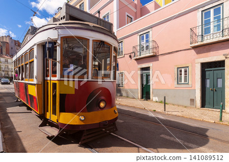 Classic yellow tram traveling through a historic street in Lisbon, Portugal, surrounded by traditional residential buildings and urban architecture. Classic yellow tram traveling through a historic street in Lisbon, Portugal, surrounded by traditional residential buildings and urban architecture. 141089512
