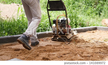 Worker legs beside plate compactor on sandy ground along concrete edging, with lush green vegetation in background, showing site preparation, soil compaction, and groundwork for Worker legs beside plate compactor on sandy ground along concrete edging, with lush green vegetation in background, showing site preparation, soil compaction, and groundwork for 141089972