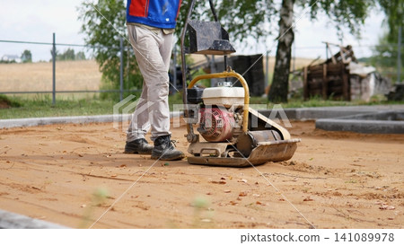 Worker in work clothes operates plate compactor on sandy ground beside fresh concrete borders, showing site preparation, soil leveling, outdoor groundwork, and rural building Worker in work clothes operates plate compactor on sandy ground beside fresh concrete borders, showing site preparation, soil leveling, outdoor groundwork, and rural building 141089978