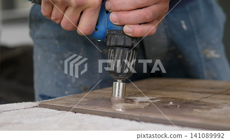 Close view of hands holding blue power drill with sanding attachment pressing into wooden board, showing workshop task, dust, metal tool parts, worn clothing, and textured work 141089992