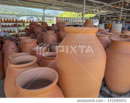 Visitors look at various large clay amphoras and terracotta pots in a pottery market outside. The market features many pots in different sizes and shapes arranged in rows. Visitors look at various large clay amphoras and terracotta pots in a pottery market outside. The market features many pots in different sizes and shapes arranged in rows. 141090031
