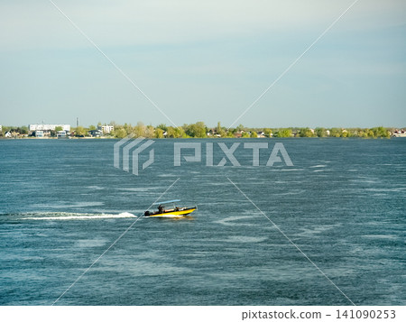 A speedboat races across open water against the backdrop of the distant Volga River shoreline under a cloudy sky A speedboat races across open water against the backdrop of the distant Volga River shoreline under a cloudy sky 141090253