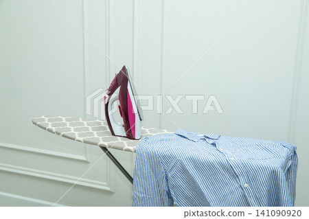 Iron rests on ironing board next to freshly washed striped shirt Iron rests on ironing board next to freshly washed striped shirt 141090920