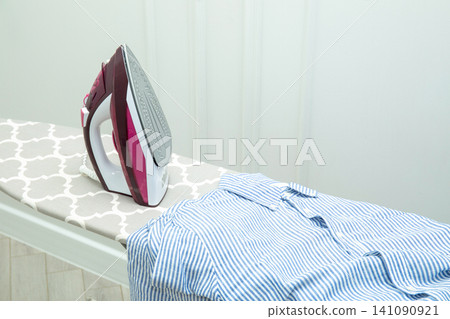 Iron rests on ironing board next to neatly arranged striped shirt Iron rests on ironing board next to neatly arranged striped shirt 141090921