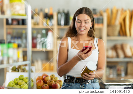 Girl with smartphone scans QR code on a jar of canned olives in the produce section of supermarket 141091420