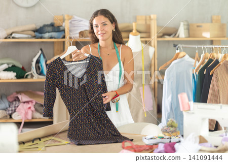 Smiling seamstress presenting handmade dress in sewing studio Smiling seamstress presenting handmade dress in sewing studio 141091444