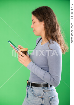 Pretty young woman scrolling through her smartphone screen against and drinking coffee green screen background. Chroma key compositing Pretty young woman scrolling through her smartphone screen against and drinking coffee green screen background. Chroma key compositing 141091712