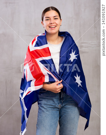 Joyful young woman wrapping herself in Australian flag while standing against gray wall indoors Joyful young woman wrapping herself in Australian flag while standing against gray wall indoors 141091927