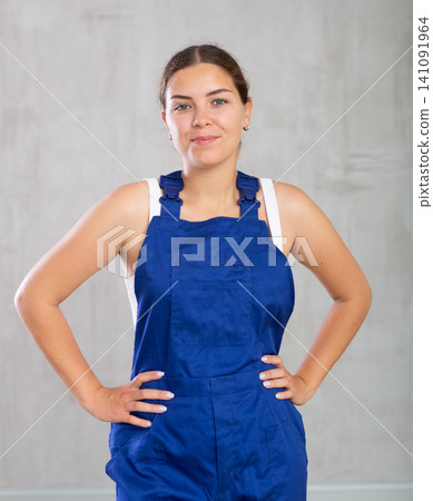 Portrait of young woman builder or mechanic in blue protective overalls on gray background Portrait of young woman builder or mechanic in blue protective overalls on gray background 141091964