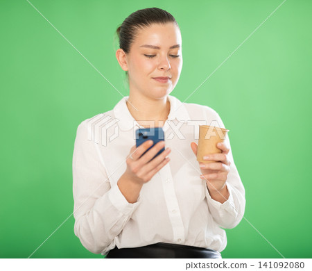 Woman drinking a hot drink while scrolling social networks on a green background Woman drinking a hot drink while scrolling social networks on a green background 141092080