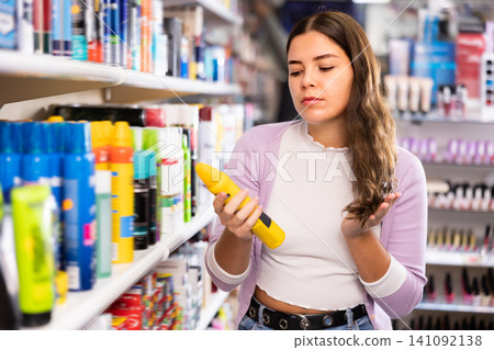 Thoughtful woman standing near shelves of hair care products and puzzled looking at her hair during choosing hairspray Thoughtful woman standing near shelves of hair care products and puzzled looking at her hair during choosing hairspray 141092138