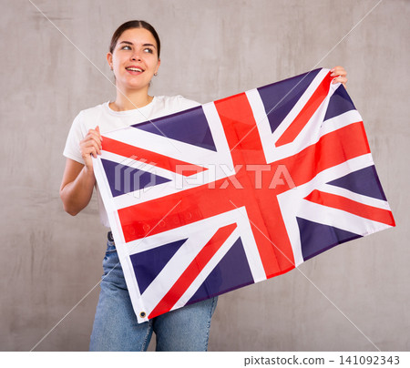 Satisfied young woman holding national flag of Great Britain on gray wall background Satisfied young woman holding national flag of Great Britain on gray wall background 141092343
