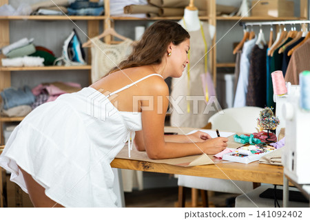 Young dressmaker drawing sketches on paper at sewing desk 141092402