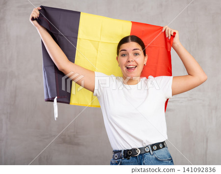 Glad young woman posing and smiling, standing with flag of Belgium against gray wall Glad young woman posing and smiling, standing with flag of Belgium against gray wall 141092836