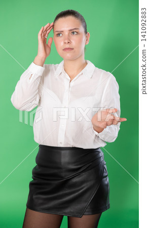 Girl in white blouse and black skirt expresses bewilderment or forgetfulness on green background Girl in white blouse and black skirt expresses bewilderment or forgetfulness on green background 141092883