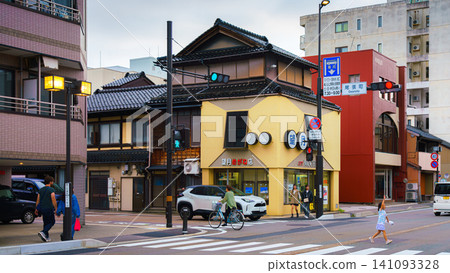 Kanazawa, Japan - Oct 6 2024, panoramic view of various buildings on a street in Kanazawa, with people crossing road in foreground and a little girl running ahead, on a cloudy day, Kanazawa, Japan 141093328