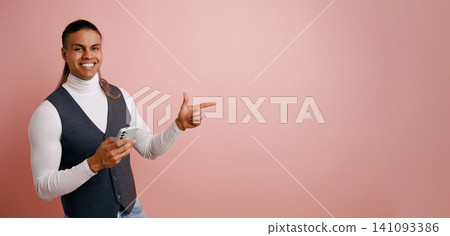 Latino man pointing while holding smartphone presenting copy space Latino man pointing while holding smartphone presenting copy space 141093386