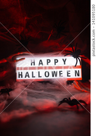 Dark still life featuring illuminated lightbox with HAPPY HALLOWEEN message holiday arrangement. Realistic skeleton arm reaches toward display, while decorative spider web creates spooky Dark still life featuring illuminated lightbox with HAPPY HALLOWEEN message holiday arrangement. Realistic skeleton arm reaches toward display, while decorative spider web creates spooky 141095180
