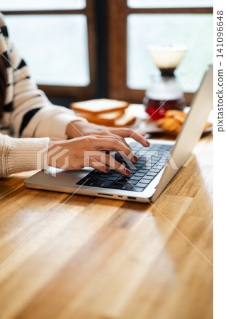 Woman Typing on Laptop at Wooden Home Desk with Coffee and Toast Breakfast in Cozy Morning Setting Woman Typing on Laptop at Wooden Home Desk with Coffee and Toast Breakfast in Cozy Morning Setting 141096648
