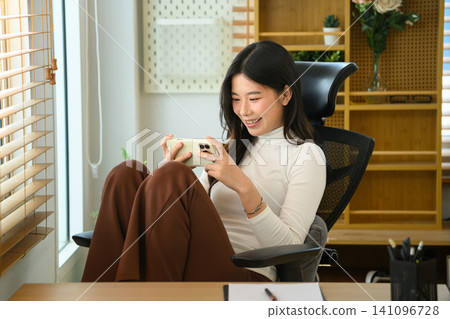 Young Asian Woman Smiling While Using Smartphone in Modern Office Chair by Window Young Asian Woman Smiling While Using Smartphone in Modern Office Chair by Window 141096728