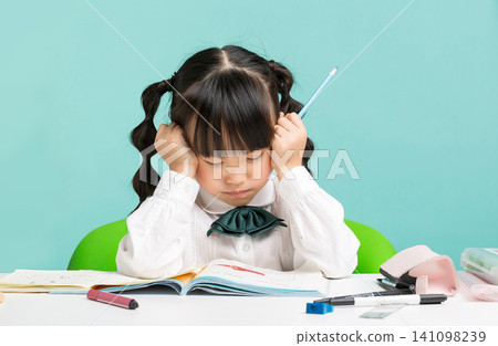 a stressed schoolgirl holding her head while writing in a notebook at her desk. a stressed schoolgirl holding her head while writing in a notebook at her desk. 141098239
