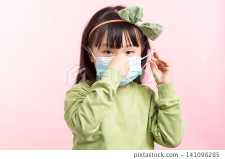 Asian little girl wearing medical face mask, adjusting mask on pink background, health protection concept Asian little girl wearing medical face mask, adjusting mask on pink background, health protection concept 141098285