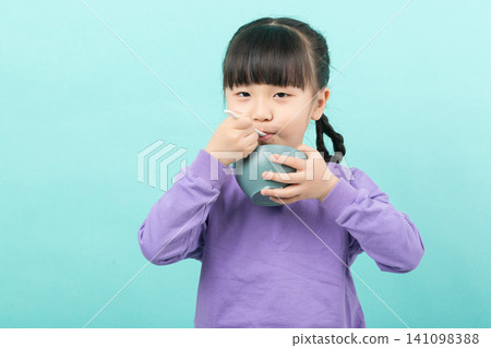Hungry little Asian girl eating food from a bowl with a spoon on a blue background. Hungry little Asian girl eating food from a bowl with a spoon on a blue background. 141098388
