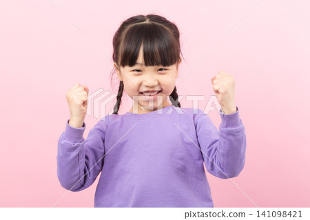 Excited little Asian girl raising fists in victory celebrating success on a pink background. 141098421
