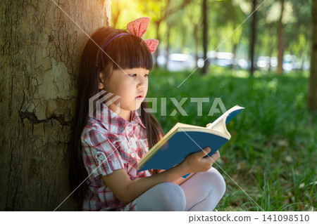 An Asian girl sits on the ground with her back against a big tree, engrossed in reading a book. An Asian girl sits on the ground with her back against a big tree, engrossed in reading a book. 141098510