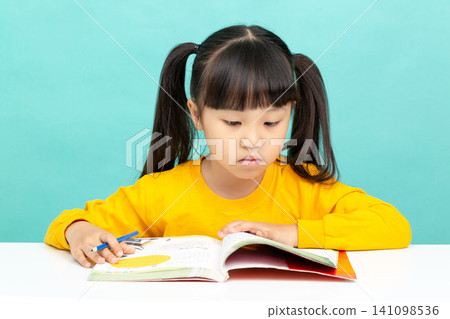 An Asian female student is reading in a blue background. Her expression is serious and focused, suggesting she may have learning difficulties. An Asian female student is reading in a blue background. Her expression is serious and focused, suggesting she may have learning difficulties. 141098536