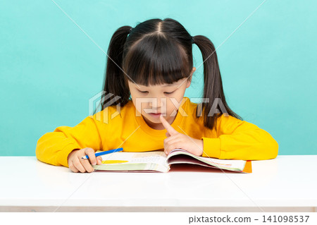 An Asian female student is reading in a blue background. Her expression is serious and focused, suggesting she may have learning difficulties. An Asian female student is reading in a blue background. Her expression is serious and focused, suggesting she may have learning difficulties. 141098537