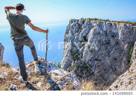 A unknown hiker(adventurer) leans on a staff and standing on the cliff is looking at vast blue ocean and mountain under big blue sky . Crimea A unknown hiker(adventurer) leans on a staff and standing on the cliff is looking at vast blue ocean and mountain under big blue sky . Crimea 141099085