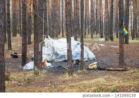 Polyethylene tent in a pine forest. Rubbish and boxes lie around the tent. Ukrainian flag on wood 141099139
