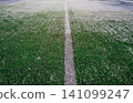 The football field with artificial green grass is covered with a light layer of snow. Early spring. Green grass on the football field is visible from under the snow. Amateur football field. 141099247