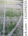 The football field with artificial green grass is covered with a light layer of snow. Early spring. Green grass on the football field is visible from under the snow. Amateur football field. 141099250