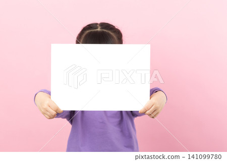 Little girl in purple shirt holding a large blank white horizontal poster with both hands, presenting copy space against a pink wall. 141099780