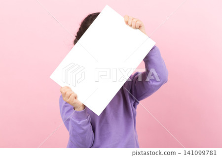 Little girl in purple shirt holding a large blank white horizontal poster with both hands, presenting copy space against a pink wall. 141099781