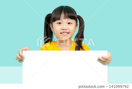 Happy little girl with pigtails holding an empty white signboard over a turquoise color background in a studio setting. Happy little girl with pigtails holding an empty white signboard over a turquoise color background in a studio setting. 141099792