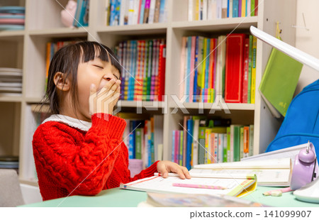 Tired little Asian girl doing homework late at night under a desk lamp, yawning and feeling exhausted from study pressure. Tired little Asian girl doing homework late at night under a desk lamp, yawning and feeling exhausted from study pressure. 141099907
