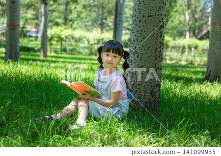 Cute elementary girl with pigtails sitting under a tree trunk in a park and holding an open book, looking at the camera. Cute elementary girl with pigtails sitting under a tree trunk in a park and holding an open book, looking at the camera. 141099935