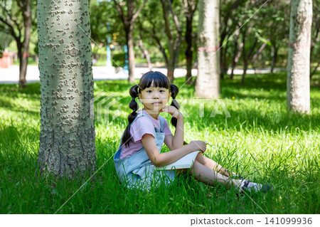 Cute elementary girl with pigtails sitting under a tree trunk in a park and holding an open book, looking at the camera. Cute elementary girl with pigtails sitting under a tree trunk in a park and holding an open book, looking at the camera. 141099936