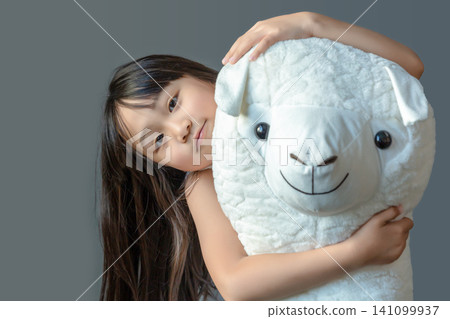 A happy young girl hugging a large white plush alpaca toy against a studio background. A happy young girl hugging a large white plush alpaca toy against a studio background. 141099937