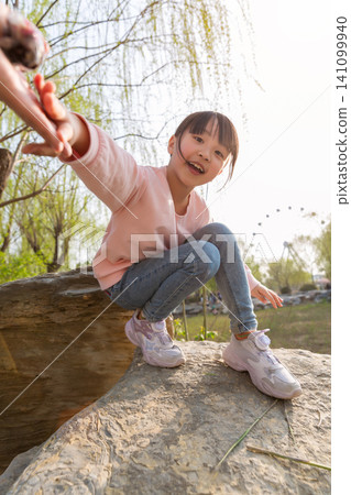 A playful young girl crouching on a rock and reaching her hand toward the camera. 141099940