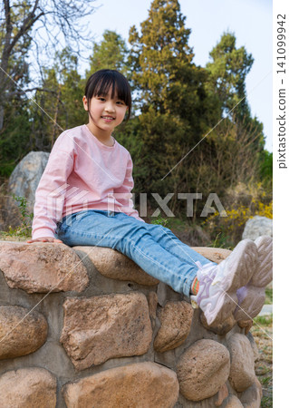Joyful little Asian girl sitting cross-legged by stone wall Joyful little Asian girl sitting cross-legged by stone wall 141099942