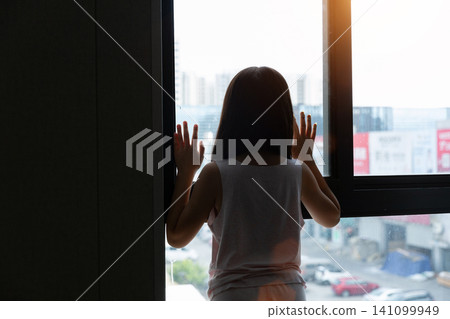 Silhouette of a lonely little girl looking out of the window at city buildings, childhood solitude and contemplation concept. Silhouette of a lonely little girl looking out of the window at city buildings, childhood solitude and contemplation concept. 141099949