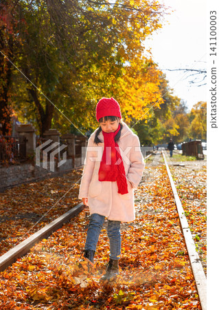 A young girl wearing a bright red hat and scarf standing on a scenic autumn railway track. A young girl wearing a bright red hat and scarf standing on a scenic autumn railway track. 141100003