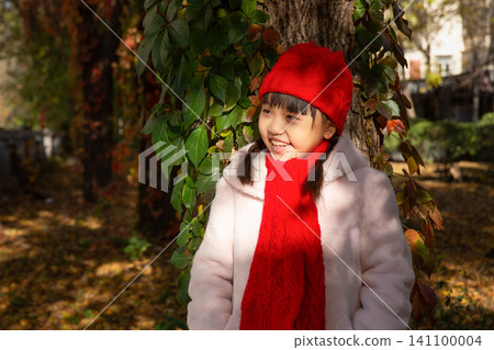 A smiling child in a red hat standing next to a tree trunk covered with green leaves in autumn. A smiling child in a red hat standing next to a tree trunk covered with green leaves in autumn. 141100004