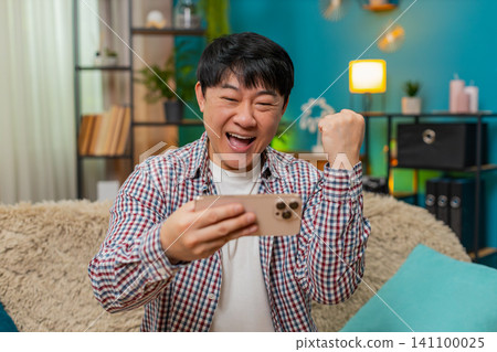 Asian man excitedly playing mobile game on smartphone, smiling loudly celebrating level victory Asian man excitedly playing mobile game on smartphone, smiling loudly celebrating level victory 141100025